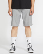 A person wearing a black shirt, Volcom Men’s Frickin Modern Stretch Shorts in light gray, white crew socks with black designs, and black-and-white sneakers stands against a plain white background.