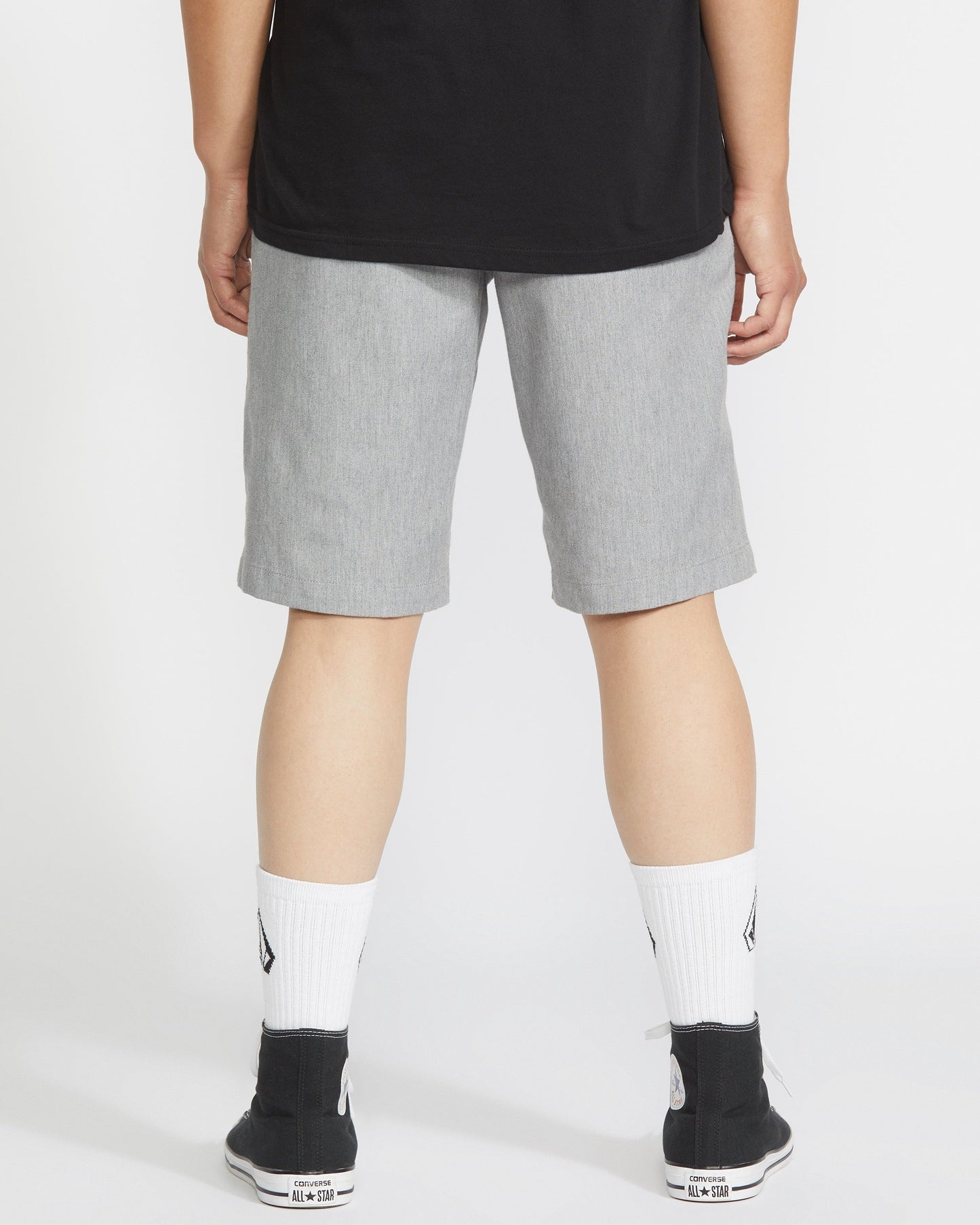 A person faces away, wearing a black shirt, Volcom Men's Frickin Modern Stretch Shorts in light gray, white socks with black logos, and black sneakers with white soles, against a plain light background.