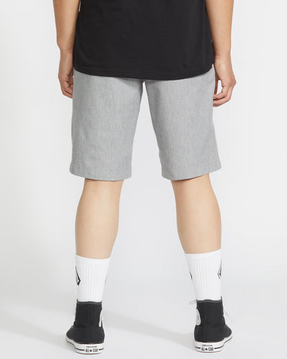 A person faces away, wearing a black shirt, Volcom Men's Frickin Modern Stretch Shorts in light gray, white socks with black logos, and black sneakers with white soles, against a plain light background.