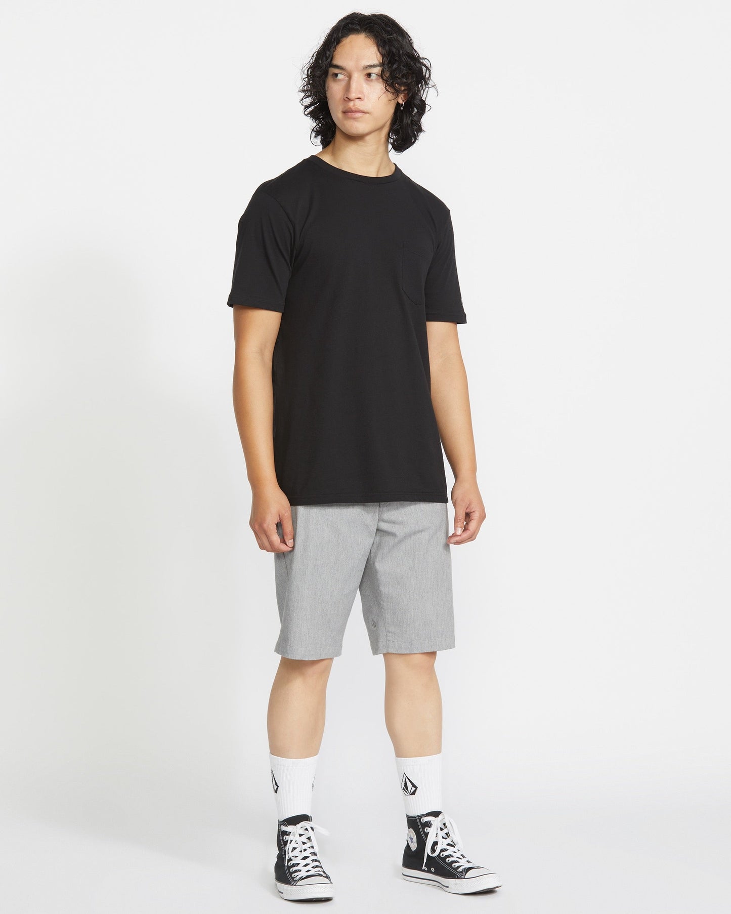 A person with medium-length curly hair wears a black t-shirt, light gray Volcom Men's Frickin Modern Stretch Shorts, white socks with black logos, and black high-top sneakers, standing against a plain white background.