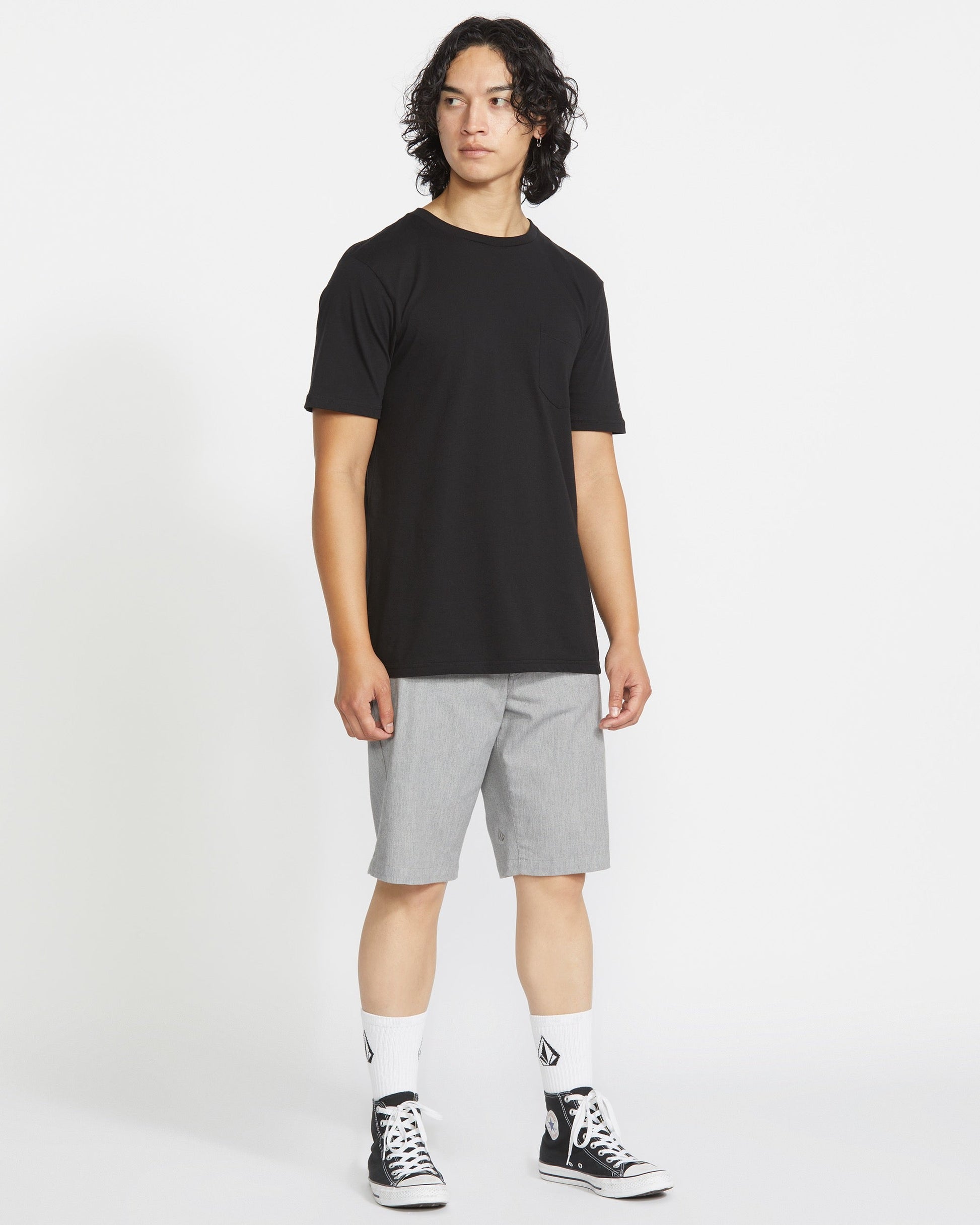 A person with medium-length curly hair wears a black t-shirt, light gray Volcom Men's Frickin Modern Stretch Shorts, white socks with black logos, and black high-top sneakers, standing against a plain white background.