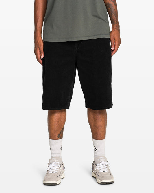 A person stands against a plain white background, showcasing their lower body in Volcom Men's Chillow 24" Shorts (antique black), paired with a green t-shirt, white socks, and white sneakers.