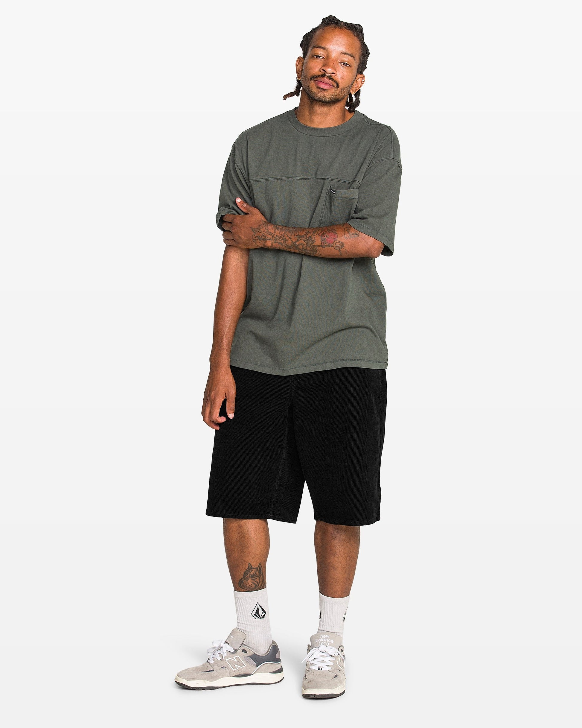 A man with braided hair stands against a white background, wearing a loose dark green tee, Volcom Men's Chillow 24" Shorts in antique black, white socks, and gray sneakers. He has one arm crossed and looks relaxed.