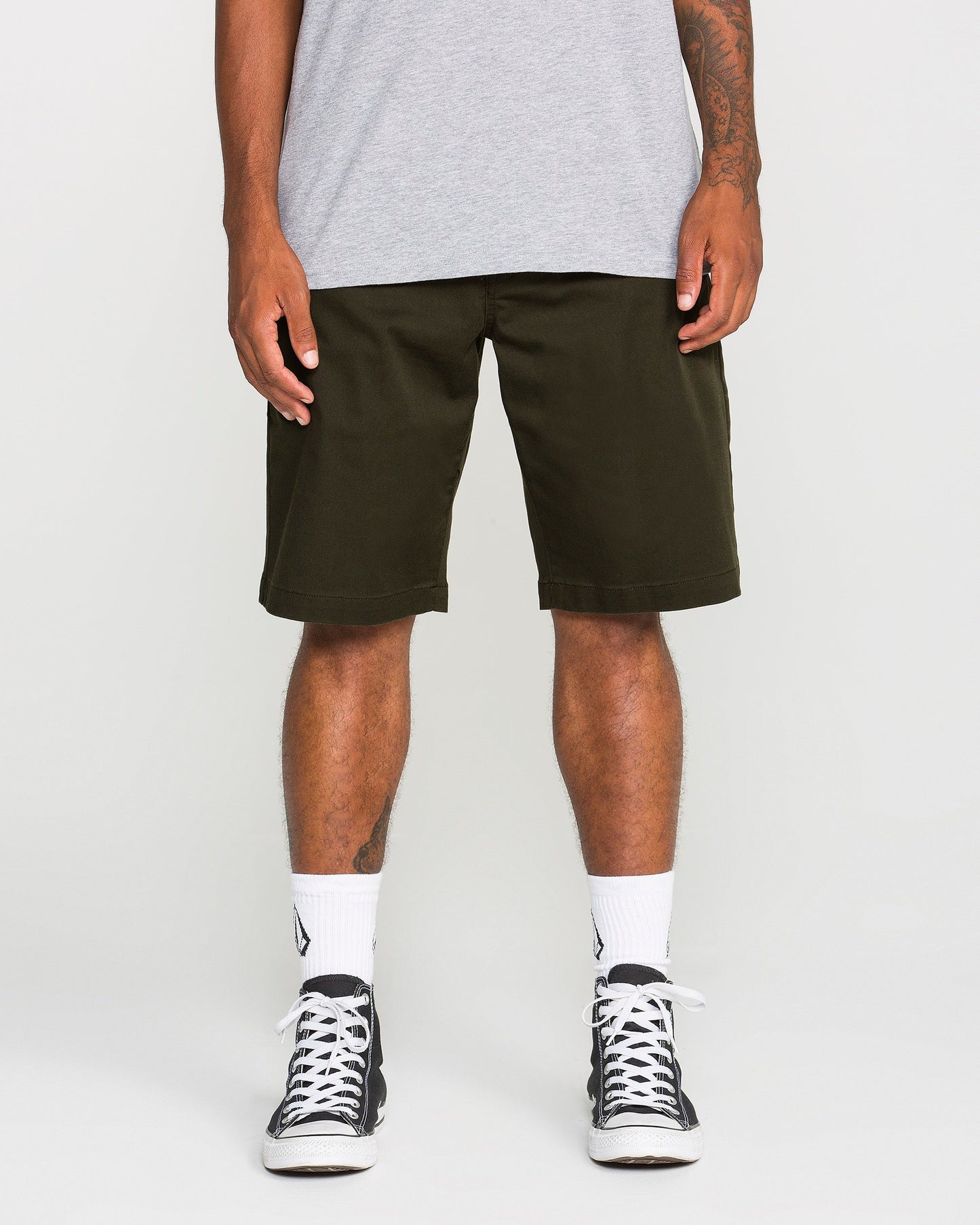 A person stands against a plain white background, shown from the shoulders down, wearing a grey t-shirt, Volcom Men's Frickin Modern Stretch Shorts in dark green—crafted from breathable cotton-poly stretch fabric—paired with white socks and black sneakers.