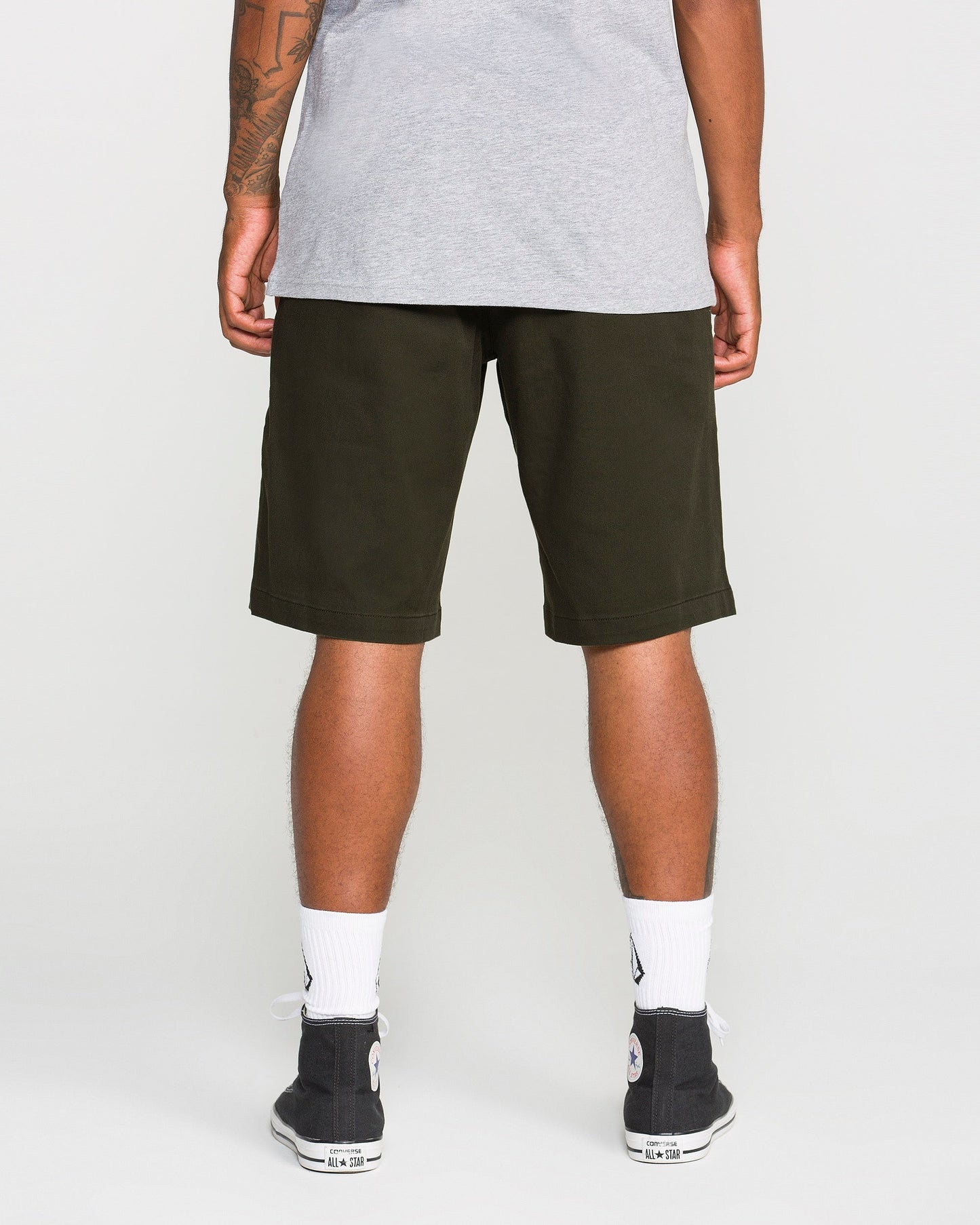 A person stands facing away, wearing a gray t-shirt, Volcom Men's Frickin Modern Stretch Shorts in dark green made from breathable cotton-poly fabric, white socks, and black sneakers.