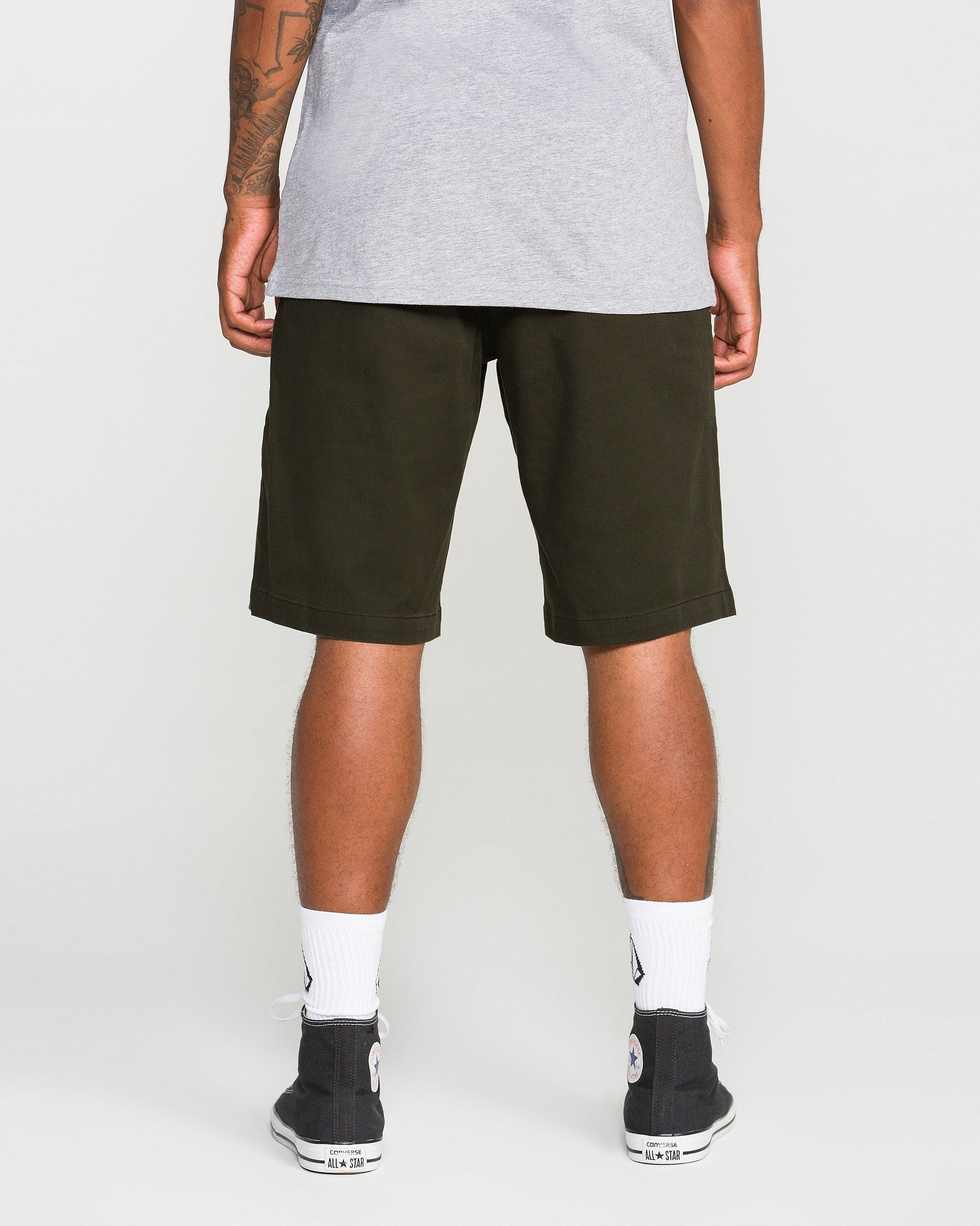 A person stands facing away, wearing a gray t-shirt, Volcom Men's Frickin Modern Stretch Shorts in dark green made from breathable cotton-poly fabric, white socks, and black sneakers.