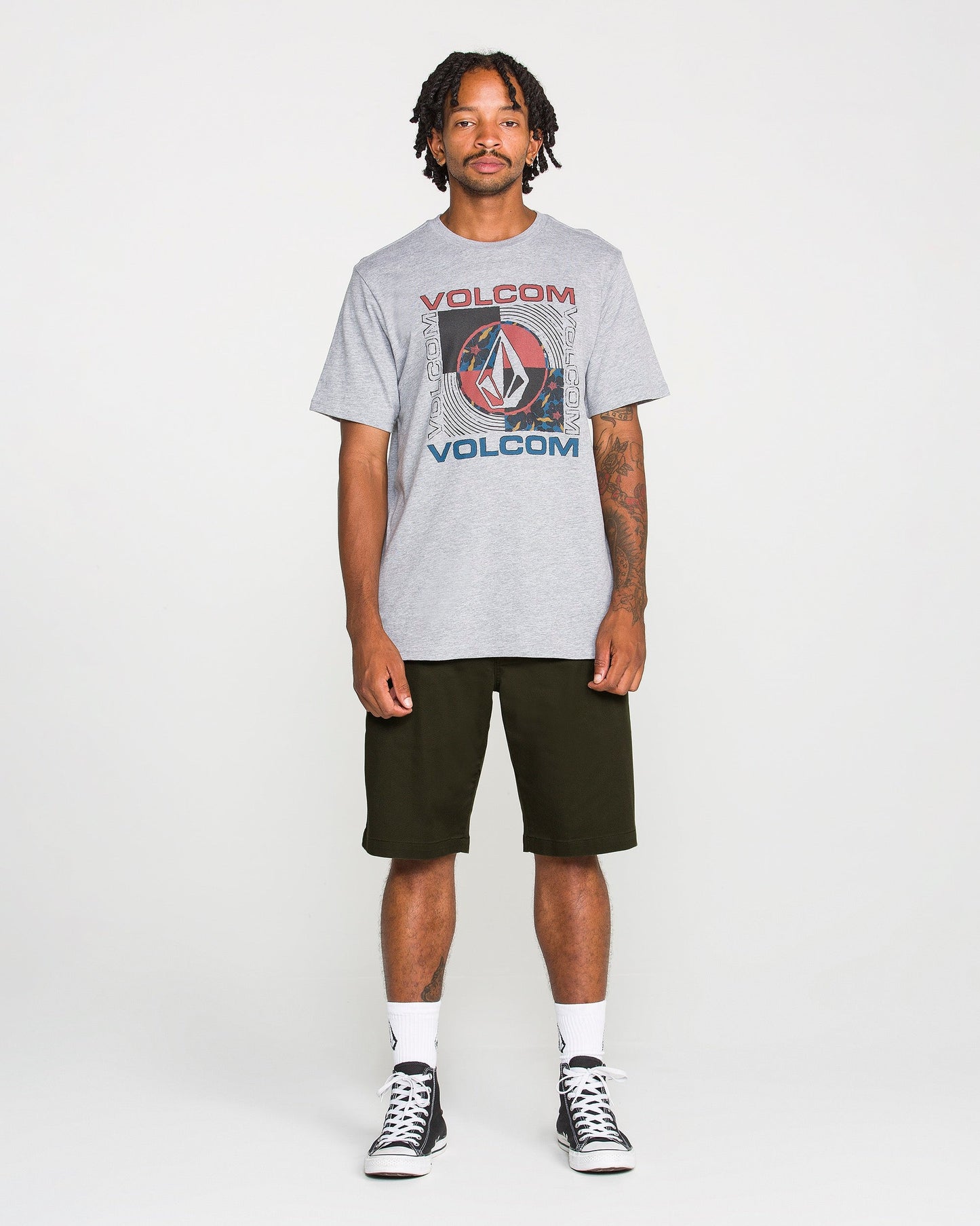 A man stands facing forward against a plain white background, wearing a gray Volcom graphic t-shirt, Volcom Men's Frickin Modern Stretch Shorts in black—crafted from breathable cotton-poly fabric—white socks, and black sneakers.