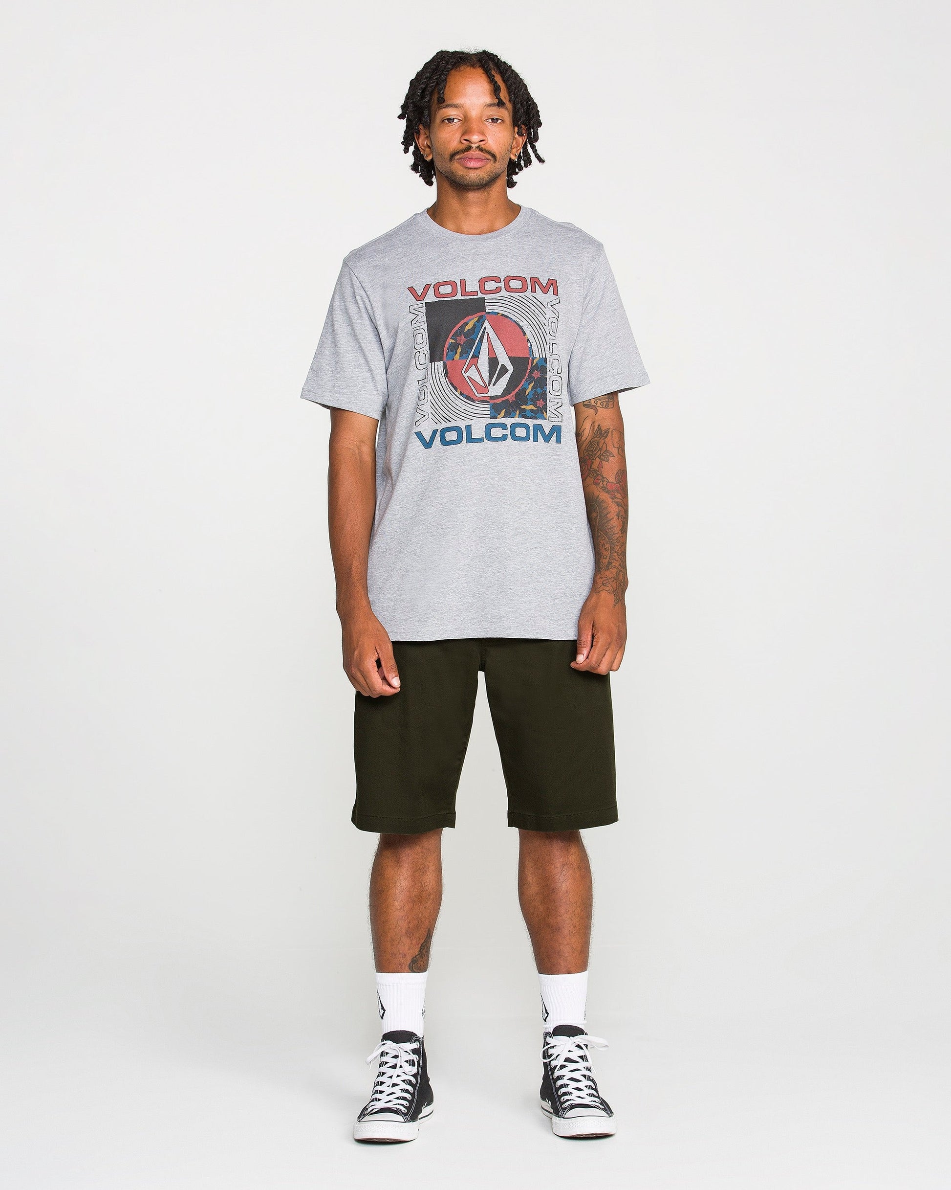 A man stands facing forward against a plain white background, wearing a gray Volcom graphic t-shirt, Volcom Men's Frickin Modern Stretch Shorts in black—crafted from breathable cotton-poly fabric—white socks, and black sneakers.