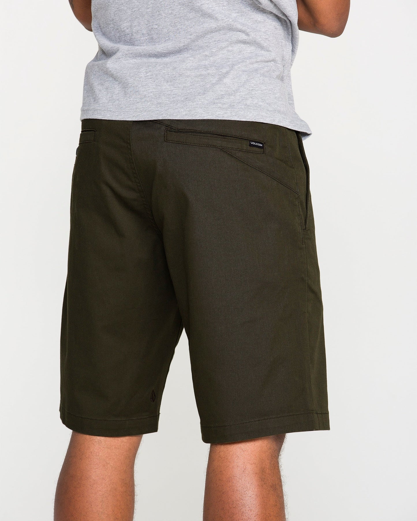 A person wearing a gray T-shirt and dark green Volcom Men's Frickin Modern Stretch Shorts is standing with their back to the camera. Only the lower torso and upper legs are visible against a plain background, emphasizing the breathable fabric of these Volcom shorts.