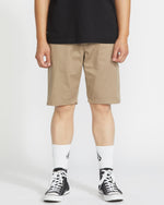A person stands against a plain white background, wearing a black t-shirt, Volcom Men's Frickin Modern Stretch 21" shorts in khaki, white crew socks with a black logo, and black low-top sneakers.