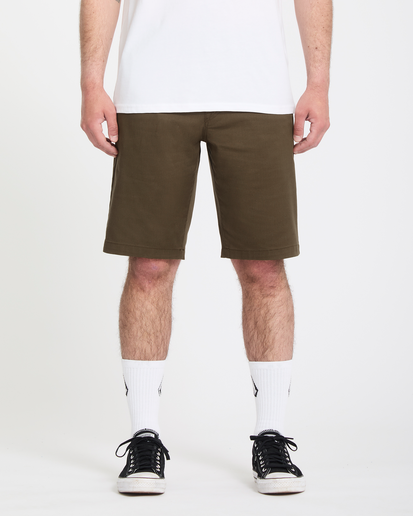 A person wearing Volcom Men's Frickin Modern Stretch Shorts in olive green, a white t-shirt, white crew socks with a logo, and black sneakers stands against a plain white background.