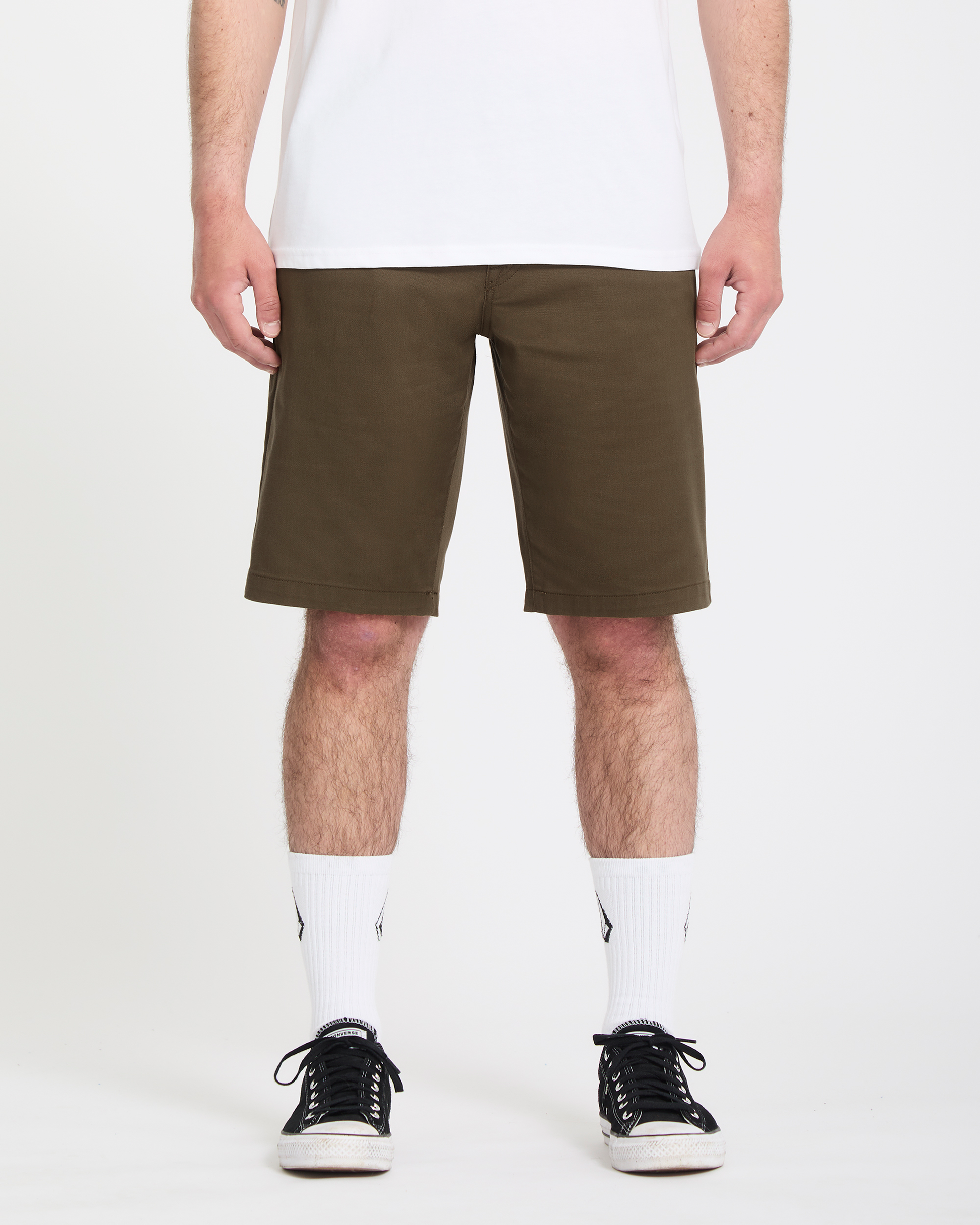A person wearing Volcom Men's Frickin Modern Stretch Shorts in olive green, a white t-shirt, white crew socks with a logo, and black sneakers stands against a plain white background.