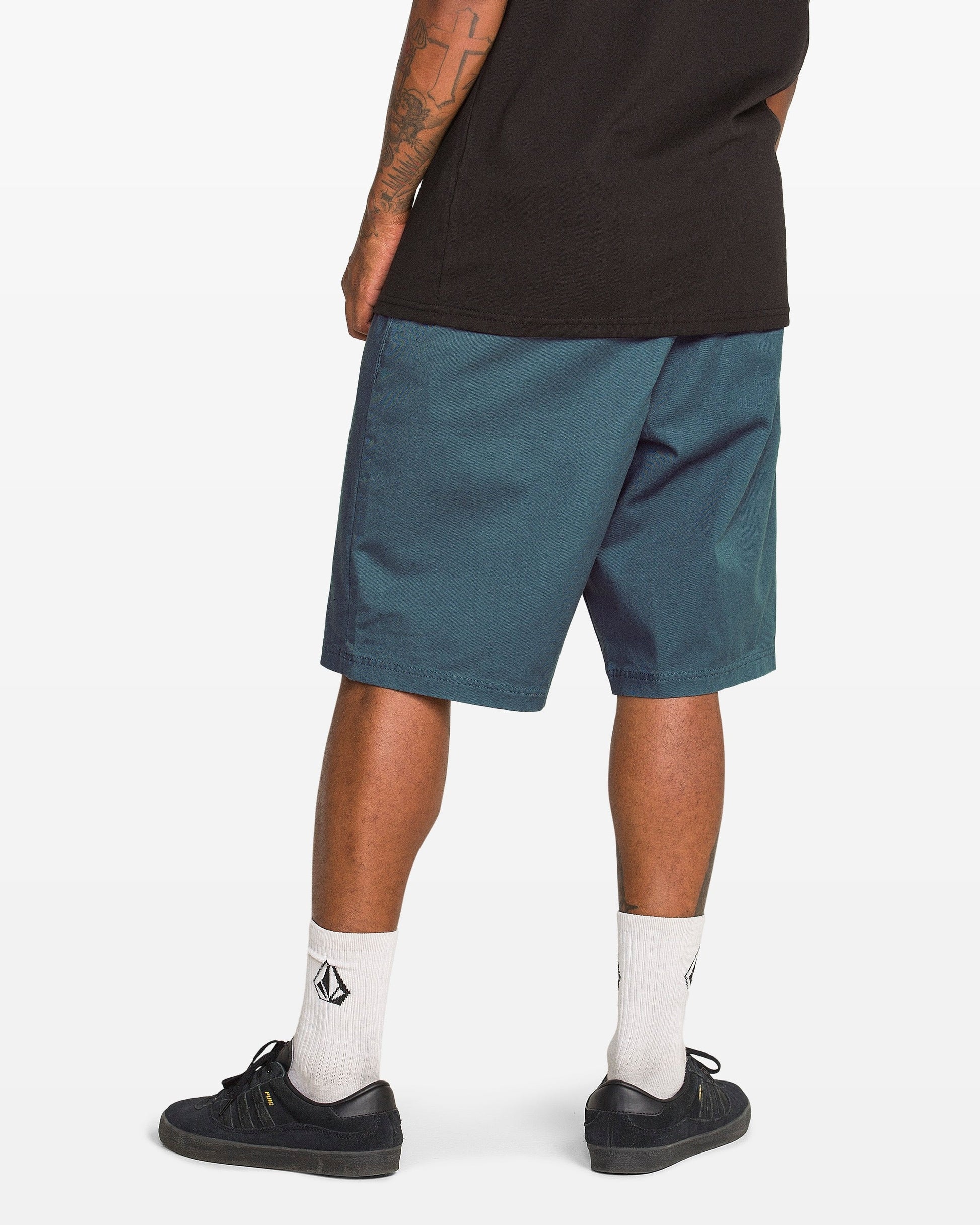 A person stands facing away, wearing a black tee, Volcom Men's Freazy Loose Elastic Waist 22" Shorts, white socks with black logos, and black sneakers. The photo highlights their legs and tattooed arms.