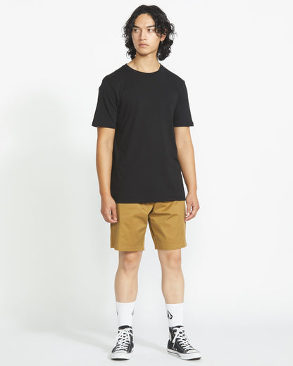 A person with medium-length curly hair wears a plain black tee, Volcom Men's Frickin Elastic Waist Shorts, white crew socks, and black high-top sneakers, standing against a white background.