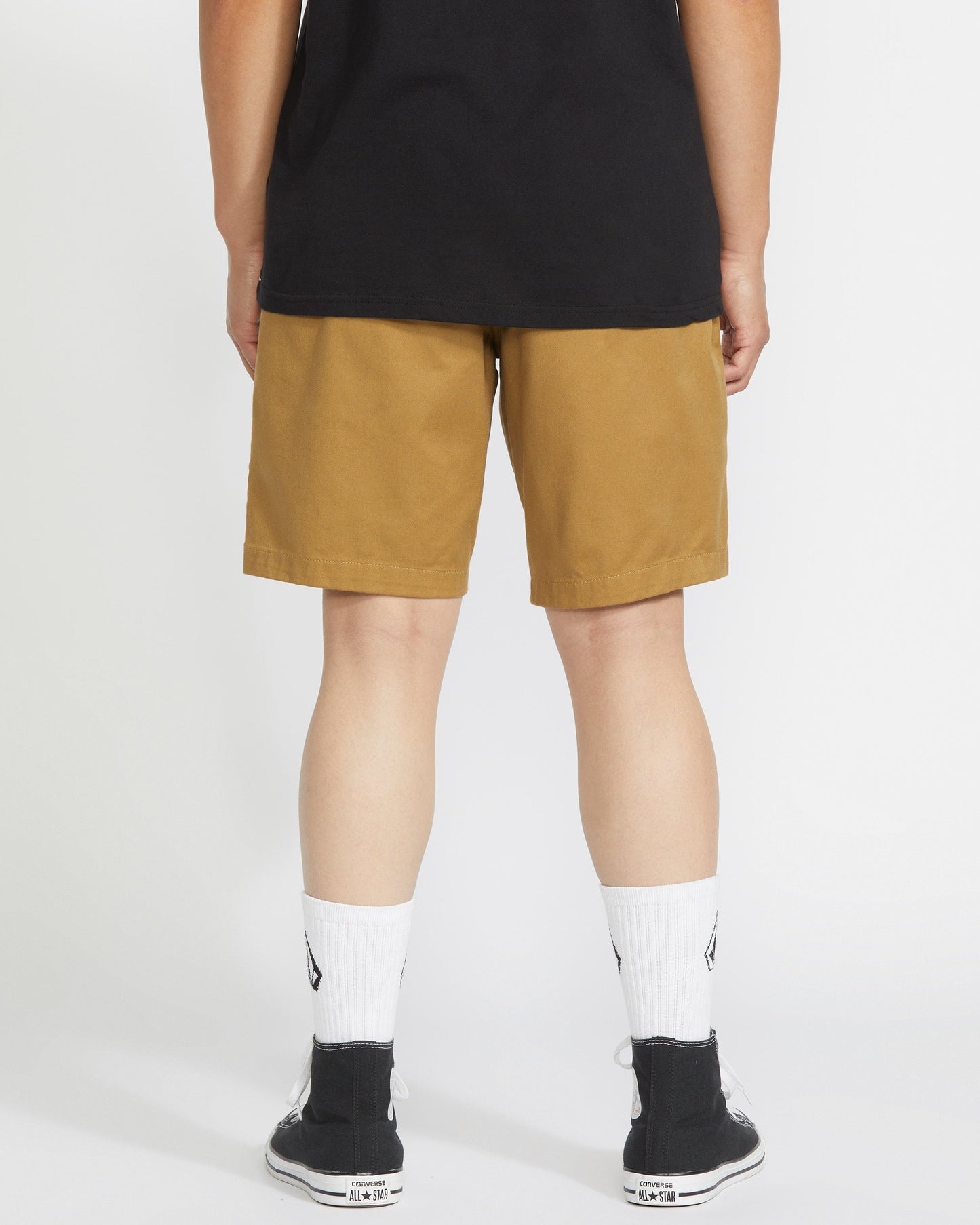 A person stands facing away from the camera wearing a black t-shirt, Volcom Men's Frickin Elastic Waist Shorts, white crew socks, and black sneakers against a plain white background.