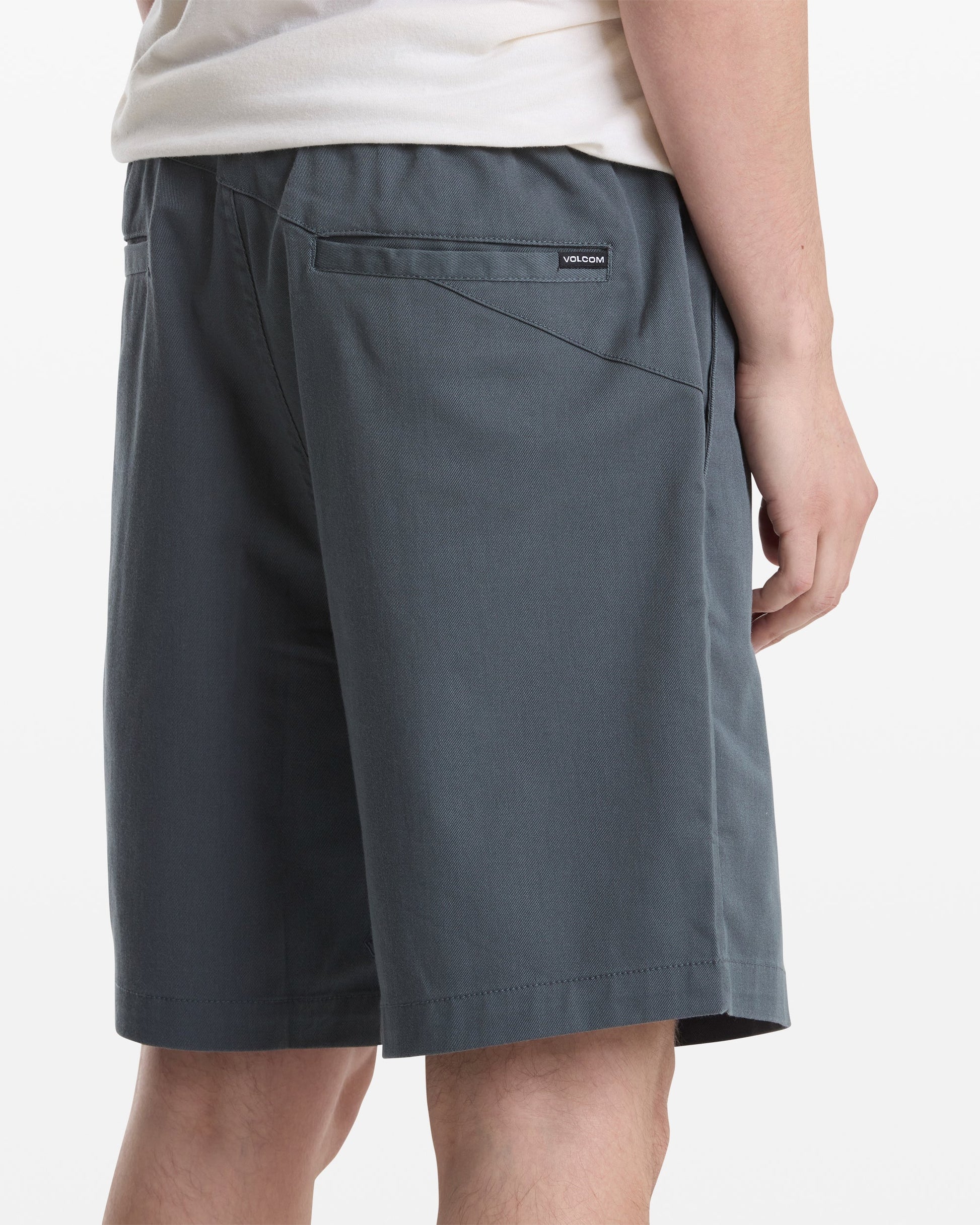A person wearing a white shirt and Volcom Men's Frickin Elastic Waist Shorts in dark gray is shown from the back and side. The shorts feature a back pocket with a small rectangular Volcom label above it.