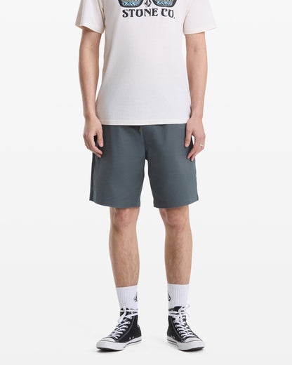 A person, head not visible, stands against a white background wearing blue-gray Volcom Men's Frickin Elastic Waist Shorts, a white graphic T-shirt, white crew socks, and black high-top sneakers.
