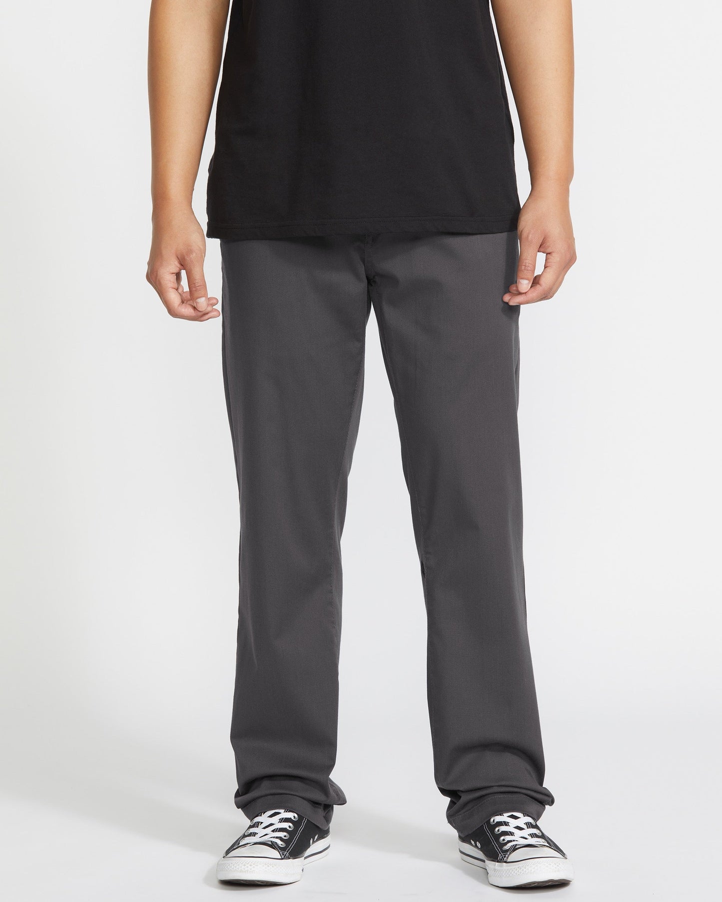 A person wears a black T-shirt, Volcom Men's Frickin Modern Stretch Pants in dark gray, and black-and-white sneakers, standing against a plain white background. The image is cropped at the shoulders.