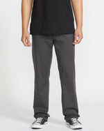 A person wears a black T-shirt, Volcom Men's Frickin Modern Stretch Pants in dark gray, and black-and-white sneakers, standing against a plain white background. The image is cropped at the shoulders.