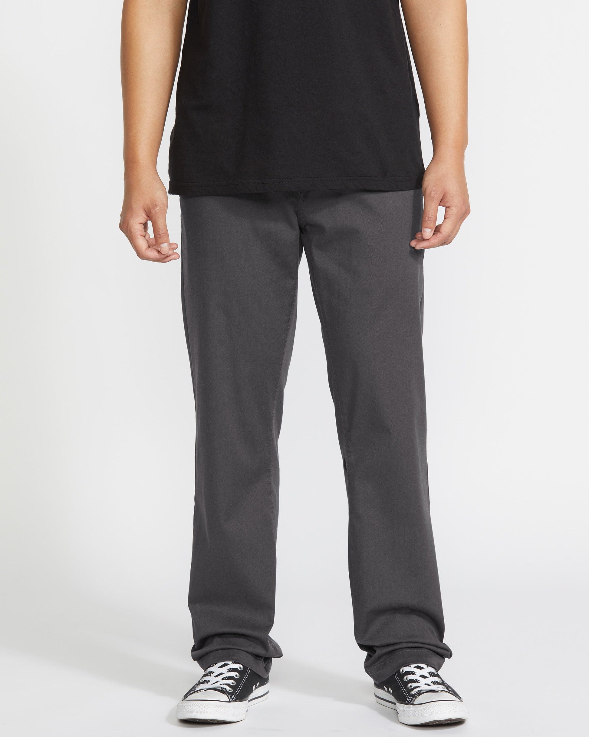 A person wears a black T-shirt, Volcom Men's Frickin Modern Stretch Pants in dark gray, and black-and-white sneakers, standing against a plain white background. The image is cropped at the shoulders.