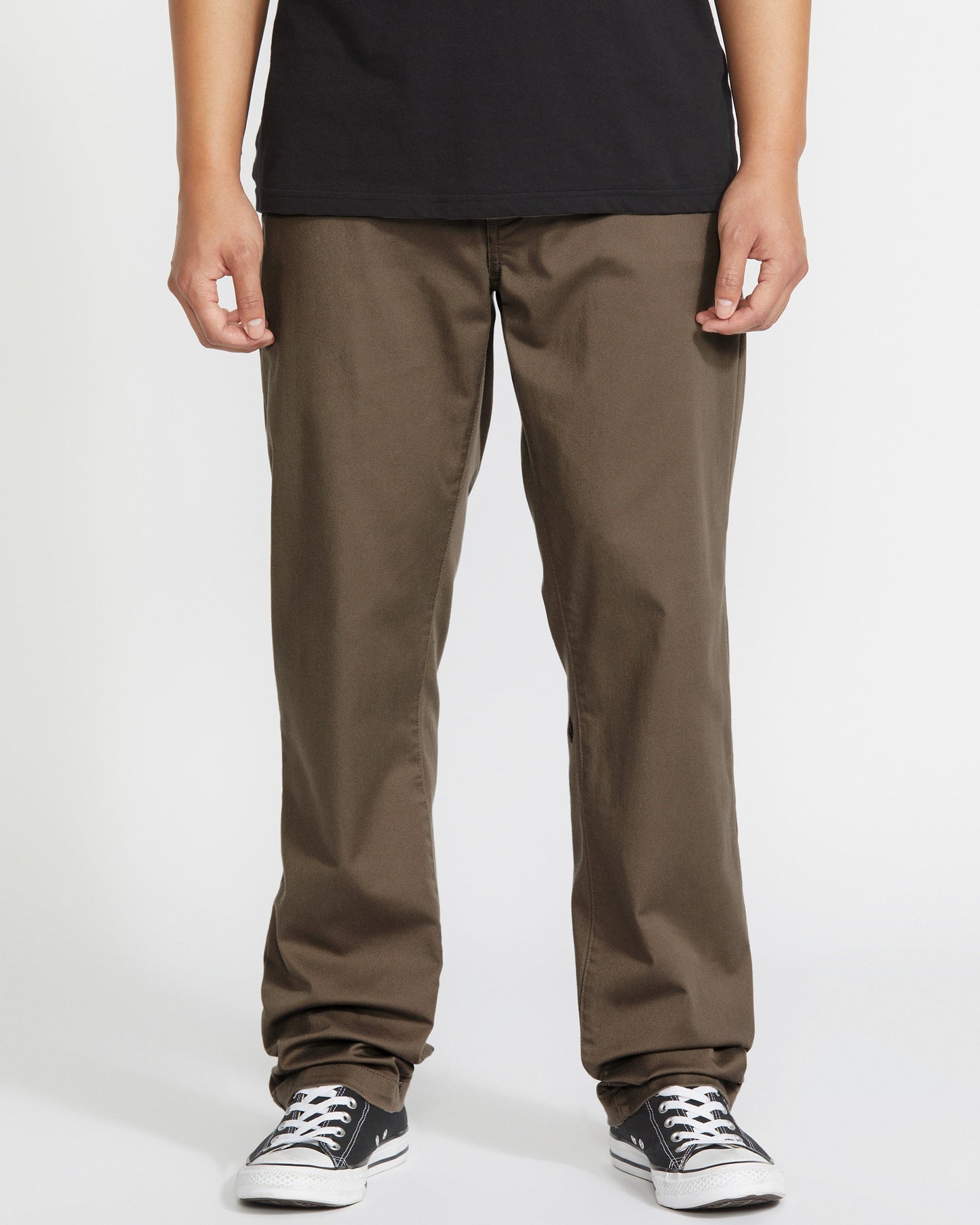 Person wearing brown pants, a black t-shirt, and black-and-white sneakers, standing against a plain white background. The photo is cropped at the shoulders, showing only the lower half of the body.
