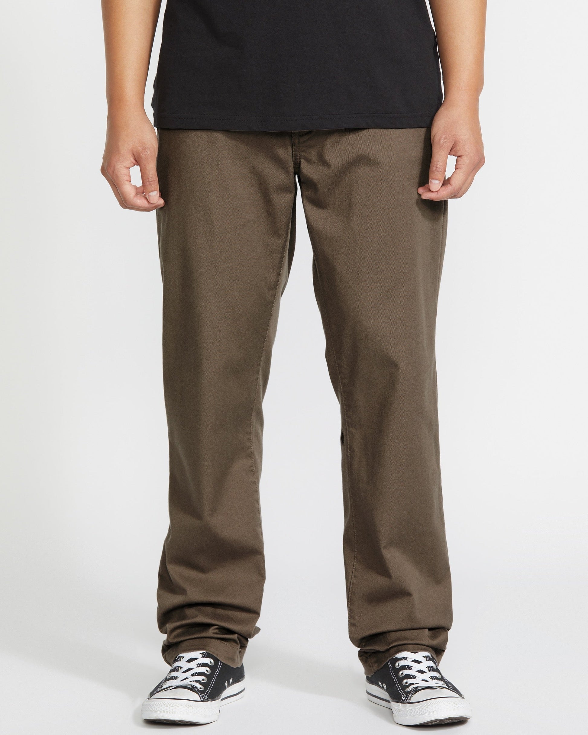 Person wearing brown pants, a black t-shirt, and black-and-white sneakers, standing against a plain white background. The photo is cropped at the shoulders, showing only the lower half of the body.