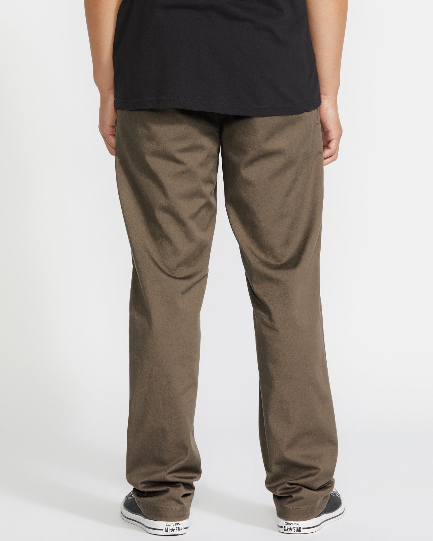 A person stands facing away from the camera, wearing a black T-shirt, brown pants, and black and white sneakers against a plain white background.
