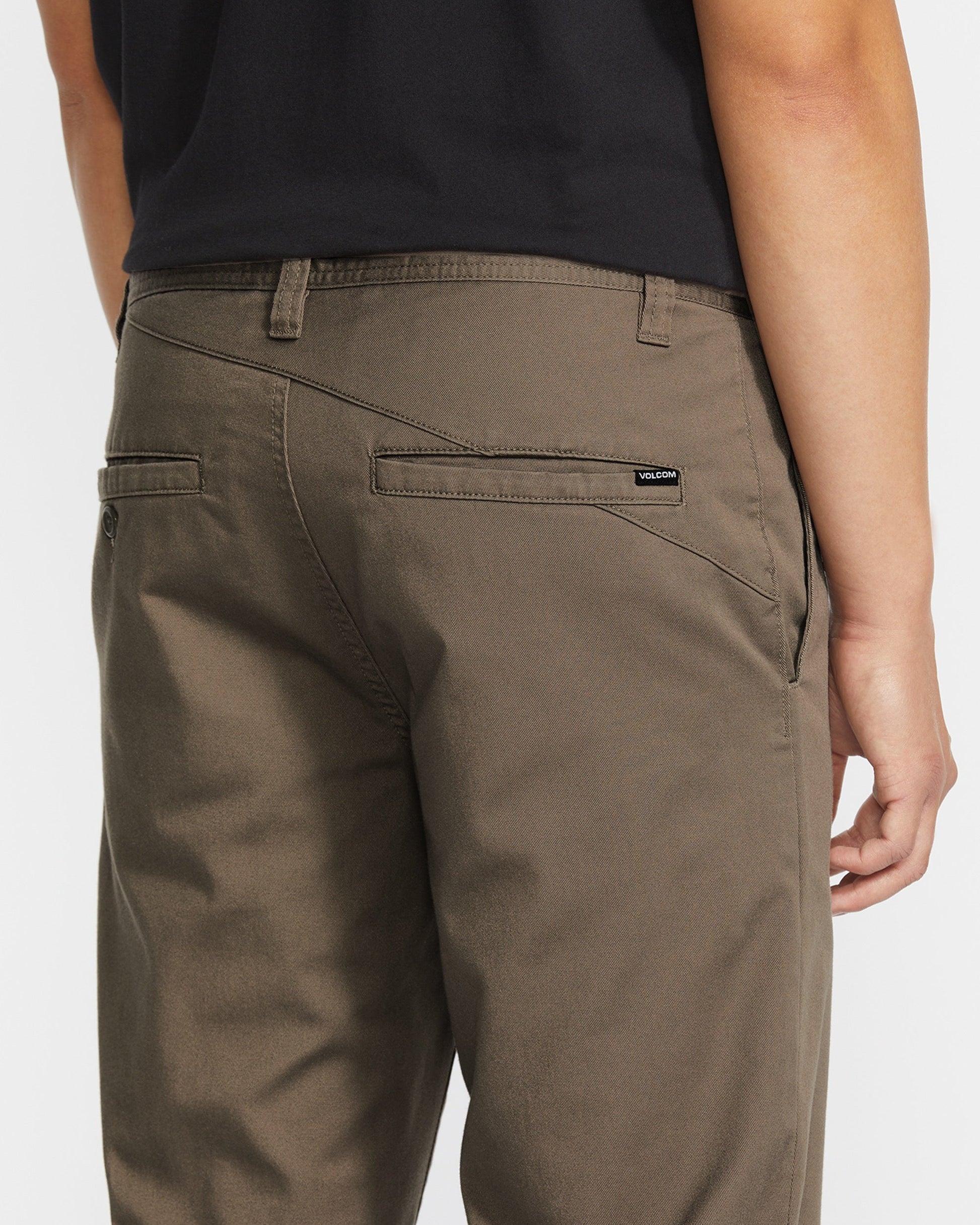 A person wearing olive green pants and a black shirt, shown from the back. The pants have two back welt pockets, one with a small black Volcom label above it.