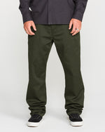 A person wearing Volcom's Men's Frickin Modern Stretch Pants in dark green, crafted with REPREVE recycled fibers, paired with a dark checkered Volcom shirt and black sneakers with white soles, standing against a plain light background.