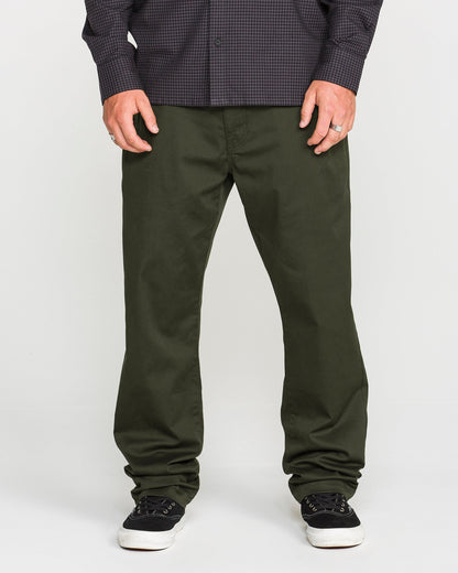A person wearing Volcom's Men's Frickin Modern Stretch Pants in dark green, crafted with REPREVE recycled fibers, paired with a dark checkered Volcom shirt and black sneakers with white soles, standing against a plain light background.