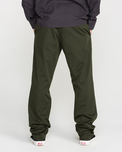 A person stands facing away, wearing Volcom Men's Frickin Modern Stretch Pants in olive green—crafted with REPREVE recycled fibers—paired with a dark long-sleeve shirt and white sneakers featuring red accents, set against a plain light background.