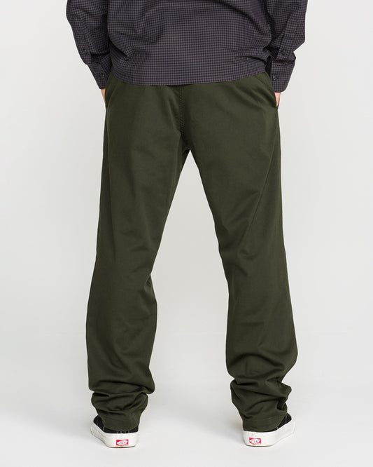 A person stands facing away, wearing Volcom Men's Frickin Modern Stretch Pants in olive green—crafted with REPREVE recycled fibers—paired with a dark long-sleeve shirt and white sneakers featuring red accents, set against a plain light background.