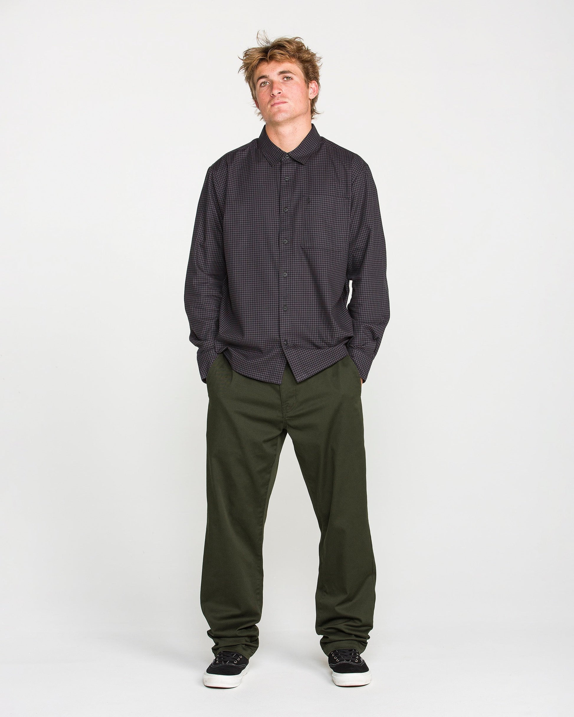 A person stands against a plain white background, wearing a dark checkered Volcom button-up shirt, Volcom Men's Frickin Modern Stretch Pants in olive green made with REPREVE recycled fibers, and black shoes, looking slightly upward with hands in pockets.