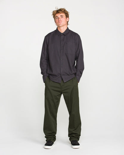 A person stands against a plain white background, wearing a dark checkered Volcom button-up shirt, Volcom Men's Frickin Modern Stretch Pants in olive green made with REPREVE recycled fibers, and black shoes, looking slightly upward with hands in pockets.