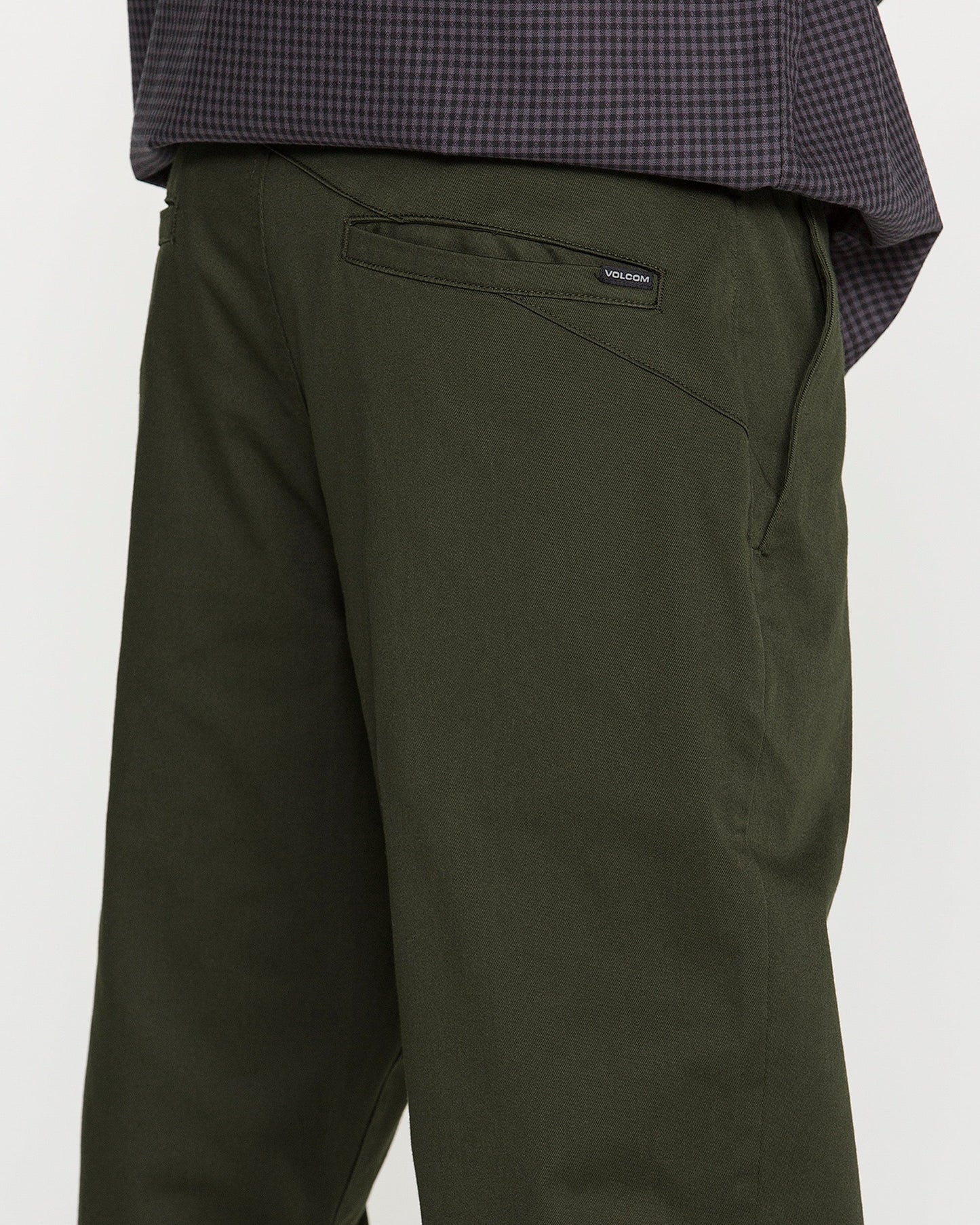 Close-up of a person wearing Volcom Men's Frickin Modern Stretch Pants in dark green, paired with a dark checkered shirt. The back pocket features a small black Volcom label, reflecting the brand’s dedication to quality. Made with REPREVE recycled fibers, these pants deliver both comfort and sustainability. The individual is standing side-on to the camera.