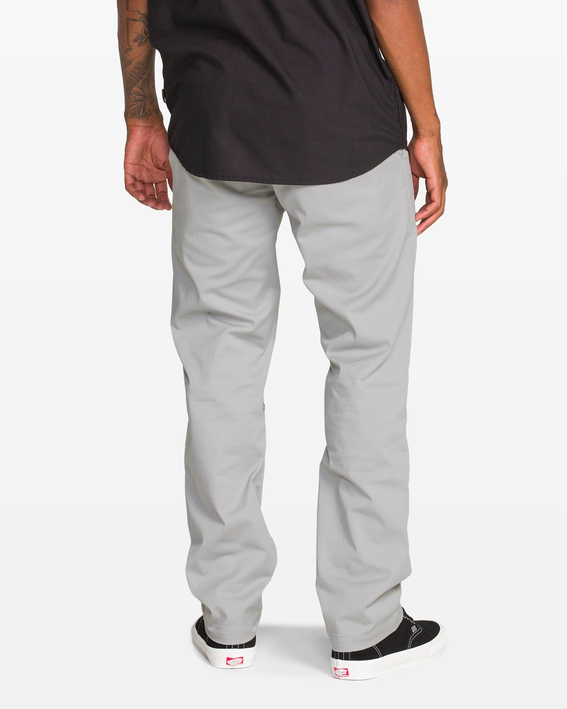 A person stands facing away, wearing light gray pants, a black shirt, and black slip-on sneakers with white soles. Only the lower back and legs are visible against a white background.