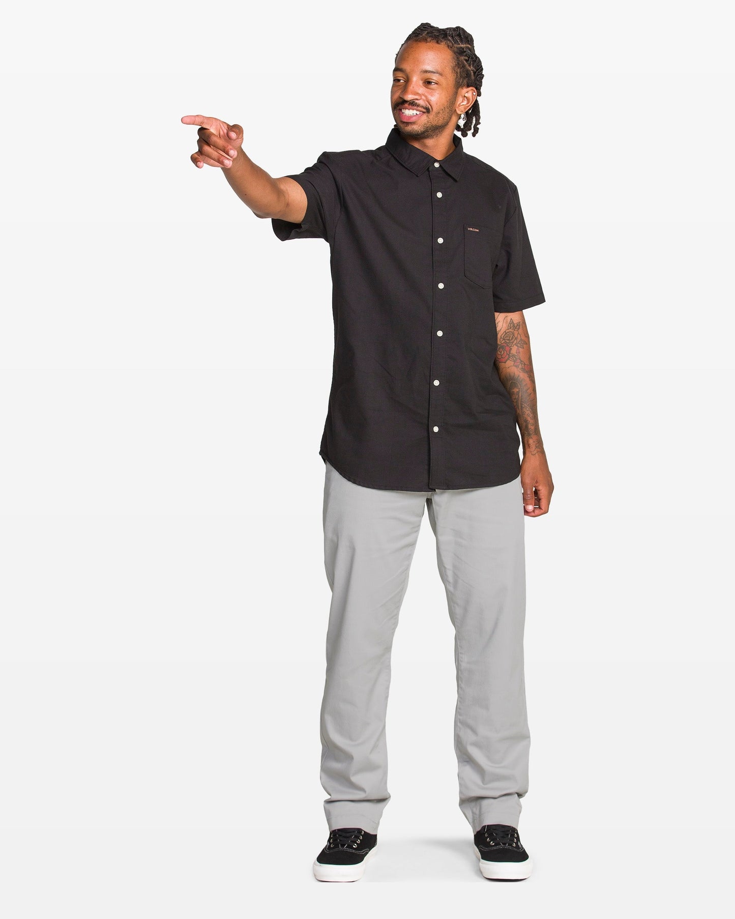 A man with braided hair wearing a black short-sleeve button-up shirt, light gray pants, and black sneakers stands smiling and pointing to his left against a plain white background.