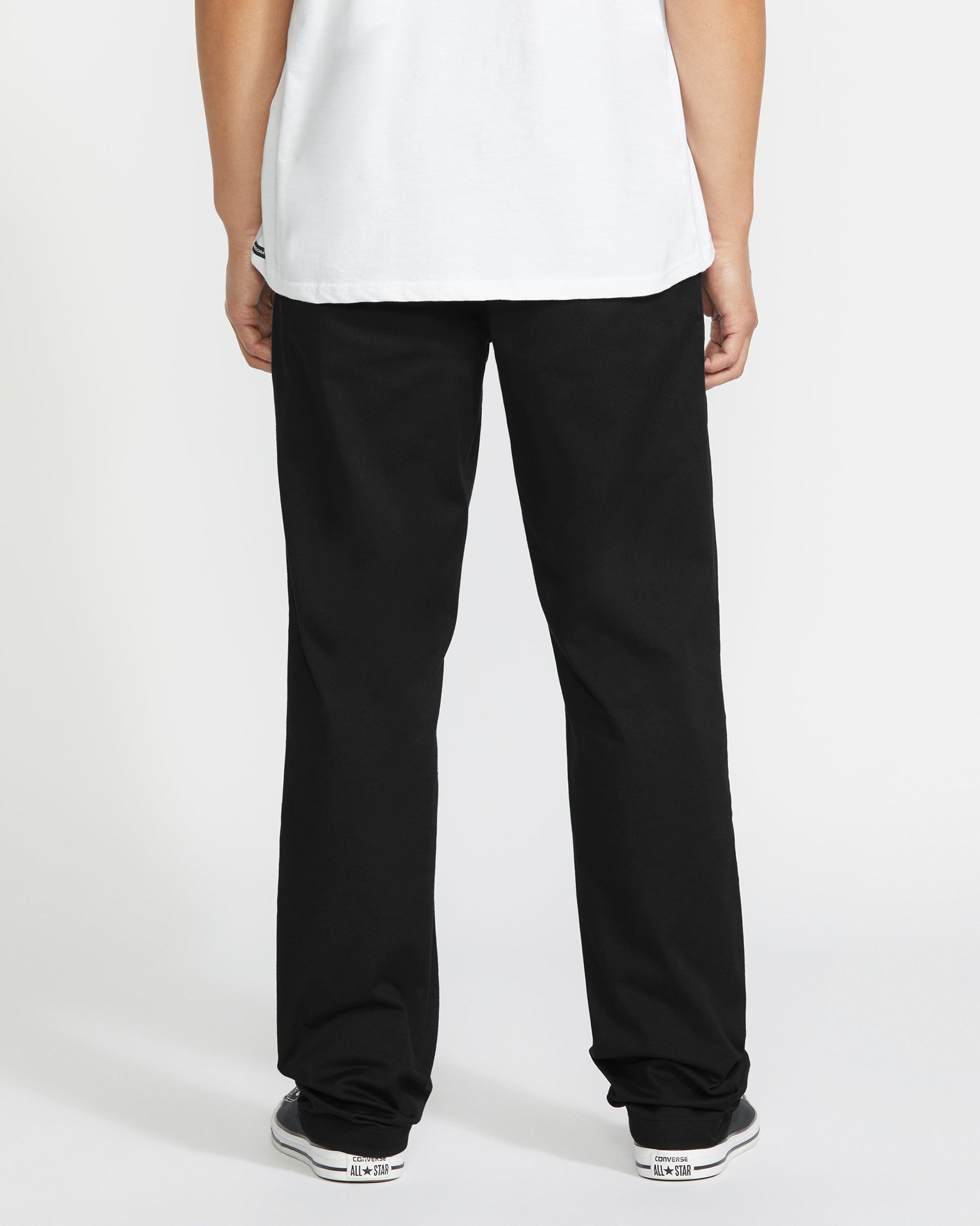 A person faces away from the camera, wearing a white t-shirt, Volcom Men's Frickin Modern Stretch Pants, and black-and-white sneakers against a plain white background.