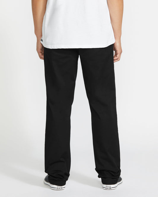 A person faces away from the camera, wearing a white t-shirt, Volcom Men's Frickin Modern Stretch Pants, and black-and-white sneakers against a plain white background.