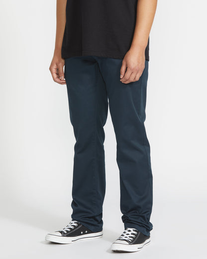A person stands against a plain white background, cropped from the shoulders down, wearing a black t-shirt, black-and-white sneakers, and Volcom Men's Frickin Modern Stretch Pants featuring slim-fit and five-pocket styling.