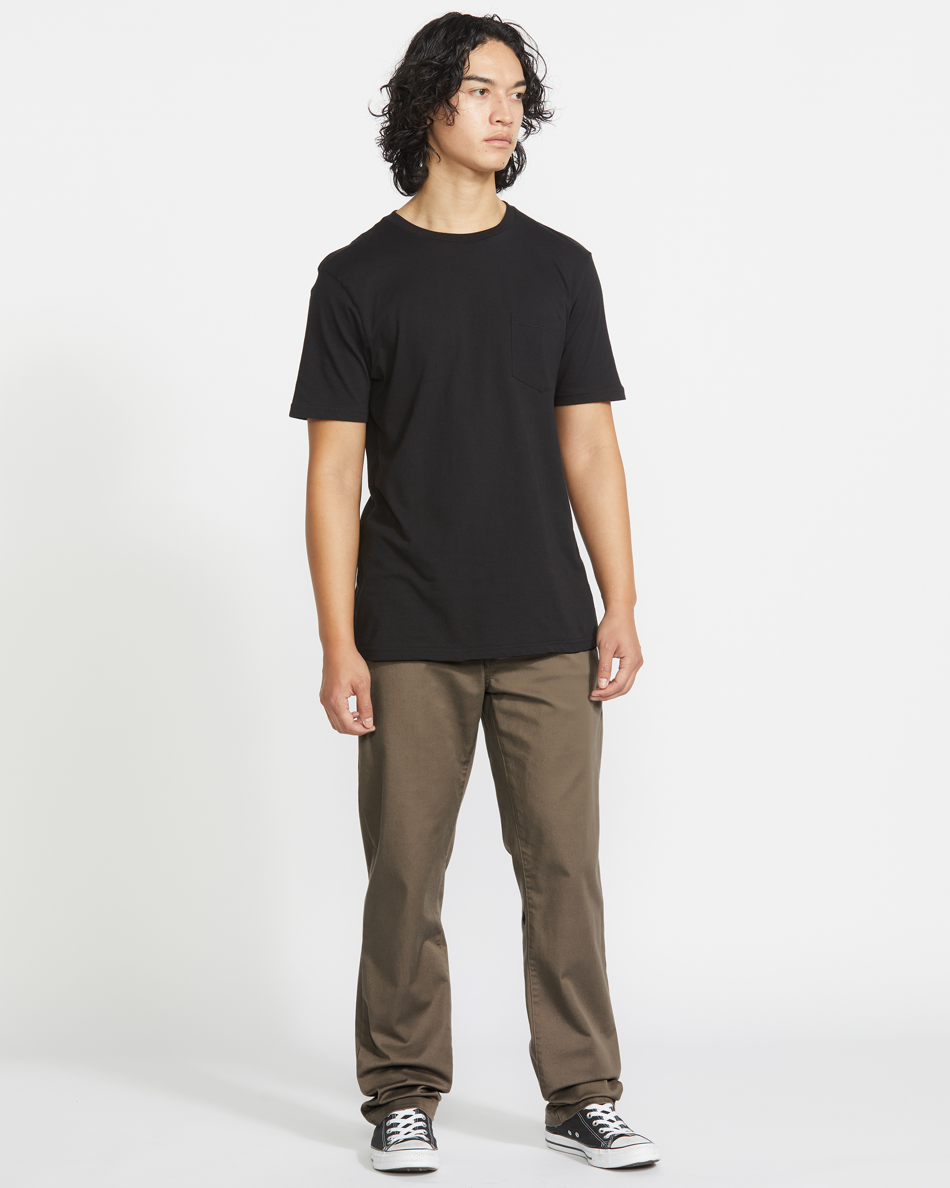 A person with shoulder-length curly hair wears a black T-shirt, Volcom Men's Frickin Modern Stretch Pants, and black sneakers, standing against a plain white background.