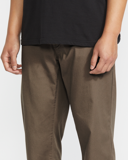 A person is shown from shoulders to knees against a white background, wearing a black shirt and brown Volcom Men's Frickin Modern Stretch Pants made with recycled polyester, standing with arms relaxed by their sides.