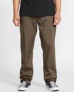 A person stands before a white background wearing Volcom Men’s Frickin Modern Stretch Pants in dark brown, made with recycled polyester, paired with black Converse sneakers.