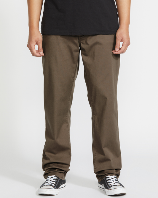 A person stands before a white background wearing Volcom Men’s Frickin Modern Stretch Pants in dark brown, made with recycled polyester, paired with black Converse sneakers.