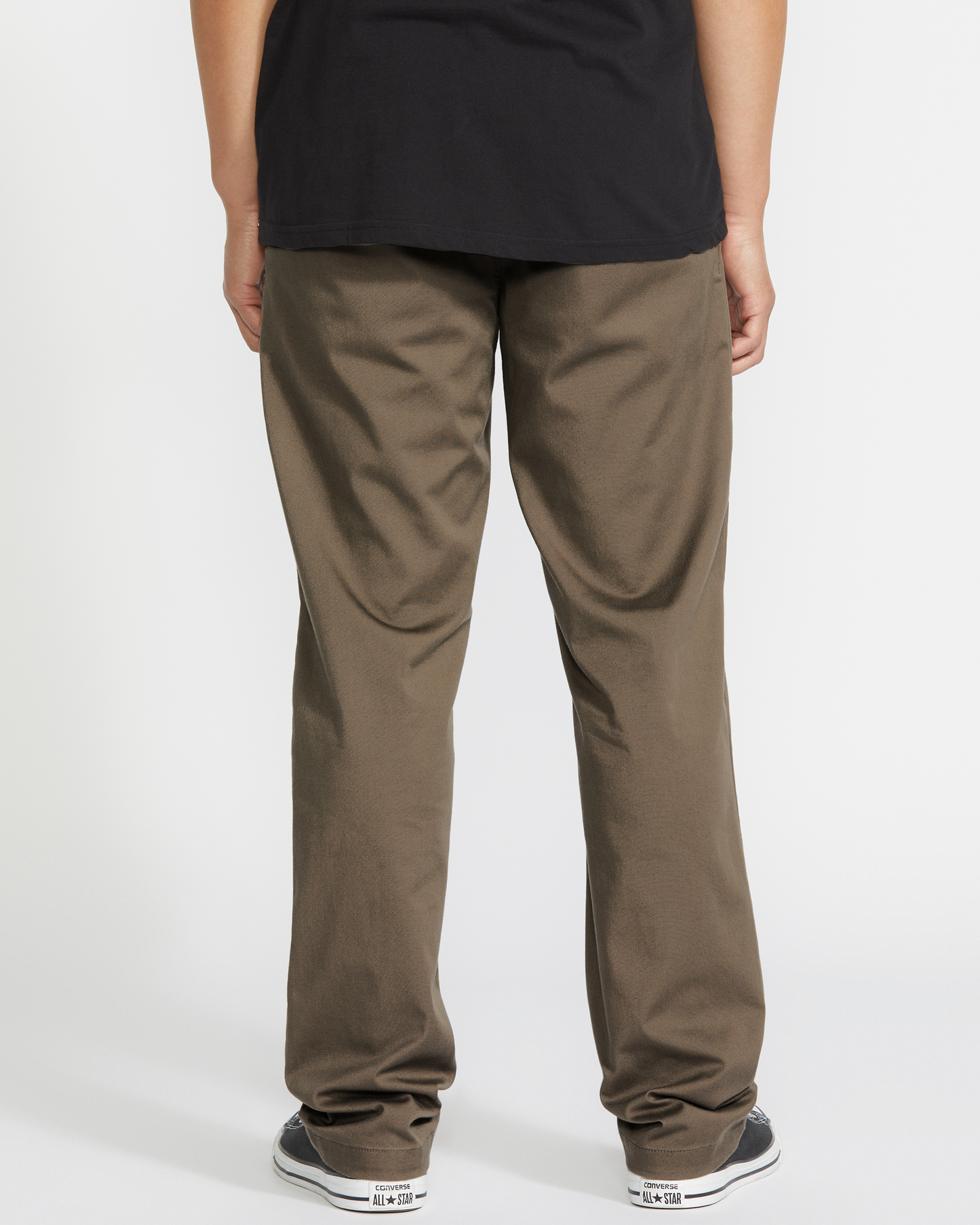 A person stands facing away from the camera, wearing a black shirt, Volcom Men's Frickin Modern Stretch Pants, and black-and-white sneakers against a plain white background.