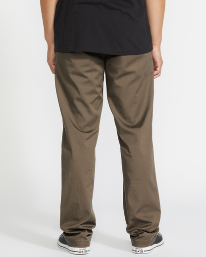 A person stands facing away from the camera, wearing a black shirt, Volcom Men's Frickin Modern Stretch Pants, and black-and-white sneakers against a plain white background.