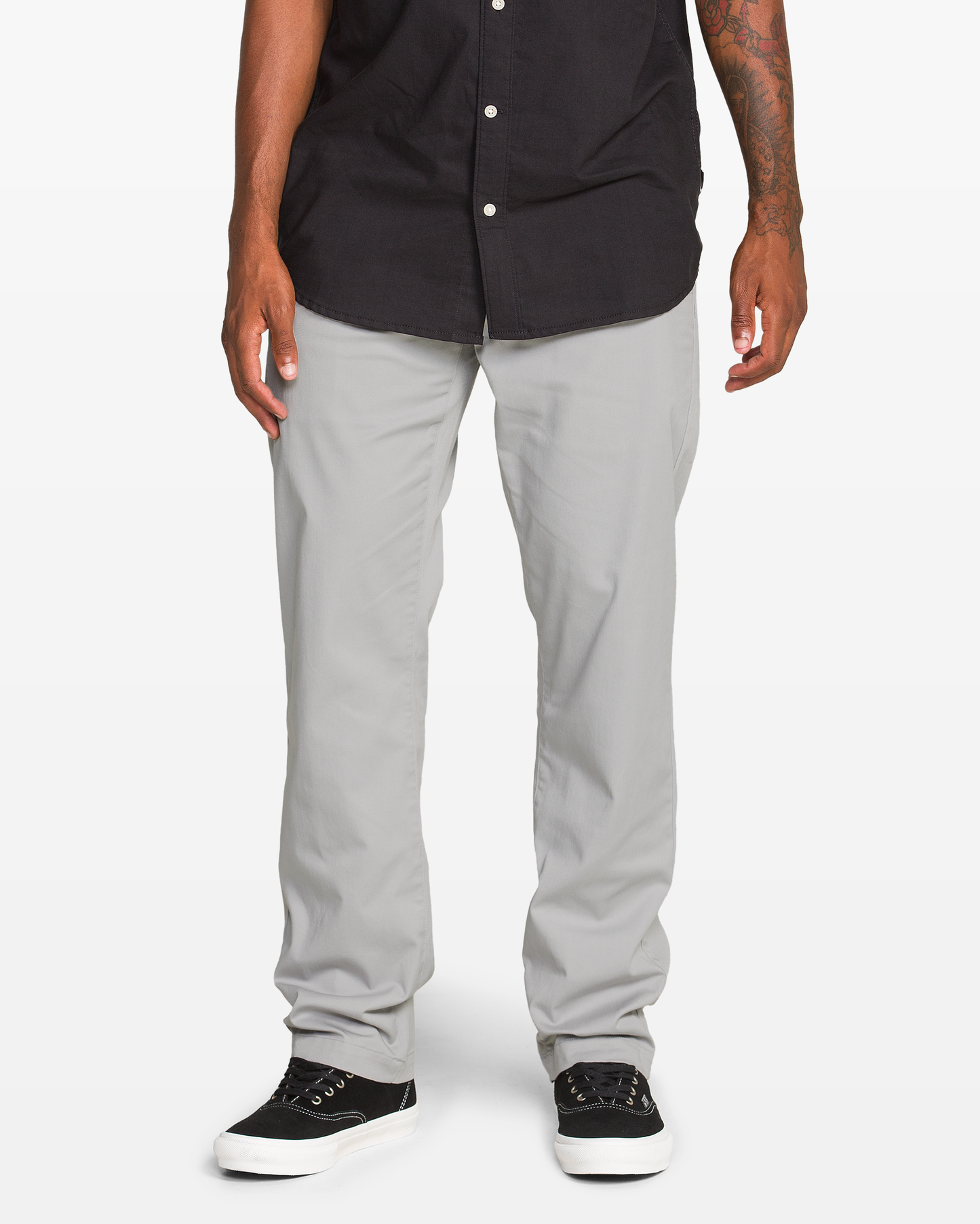 A person wearing Volcom Men's Frickin Modern Stretch Pants, a black button-up shirt, and black sneakers with white soles stands against a plain white background. Only the lower body is visible.