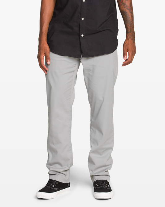 A person wearing Volcom Men's Frickin Modern Stretch Pants, a black button-up shirt, and black sneakers with white soles stands against a plain white background. Only the lower body is visible.