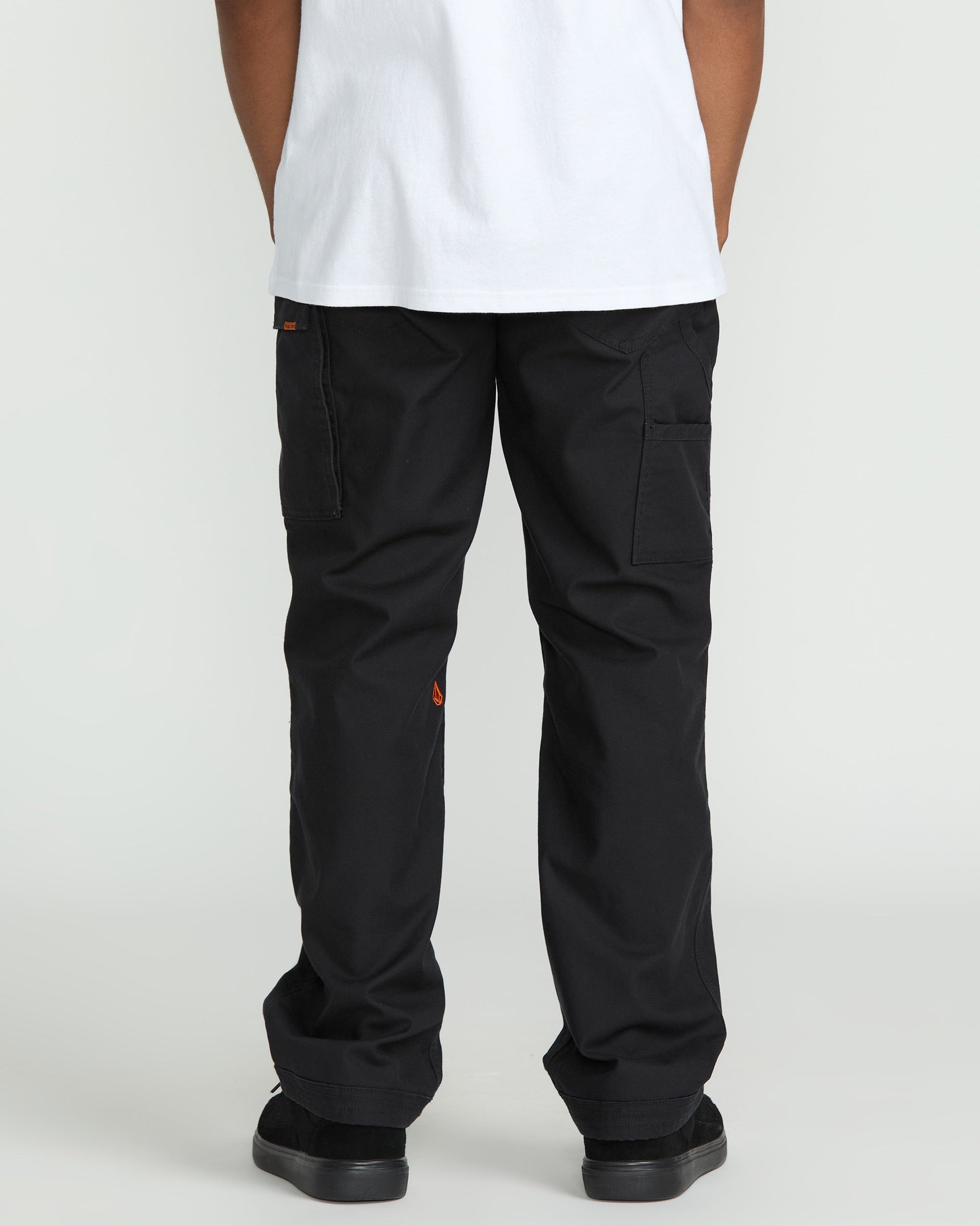 A person wearing a white T-shirt and Volcom Men's Caliper II Relaxed Work Pants in black stretch canvas with cargo pockets and small orange logo details stands facing away from the camera, paired with black shoes.