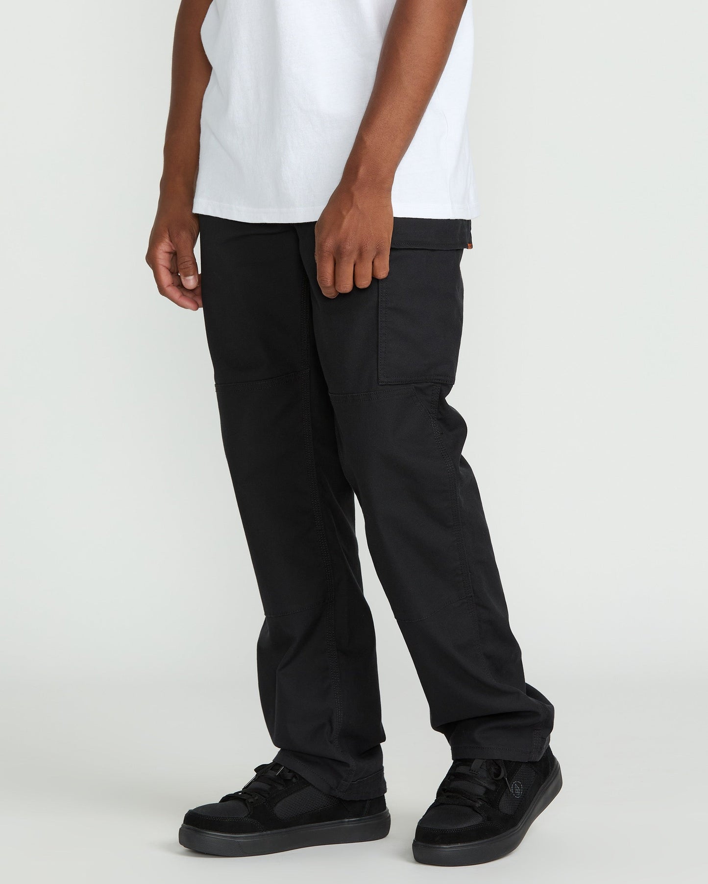 A person, visible only from the waist down, stands against a plain light background wearing Volcom Men's Caliper II Relaxed Work Pants with cargo pockets, paired with a white t-shirt and black sneakers.